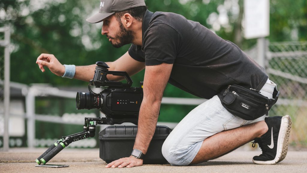 Casually dressed male videographer on hands, knees; with camera, facing left. Color.
